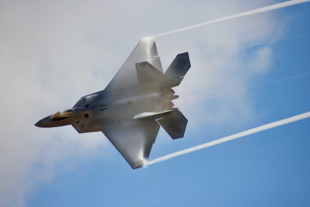 A jet fighter F-22 Raptor in flight at top speed across a blue sky with clouds. 