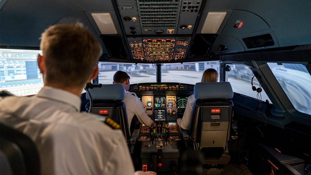 A backview of a supervisor and young people taking part in aviation training: learning to fly an aircraft using a flight simulator.