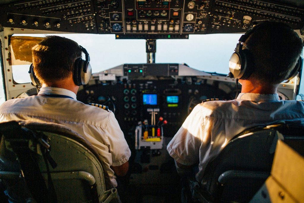 Two male pilots wearing headphones and sitting in a cockpit, looking through the windshield.