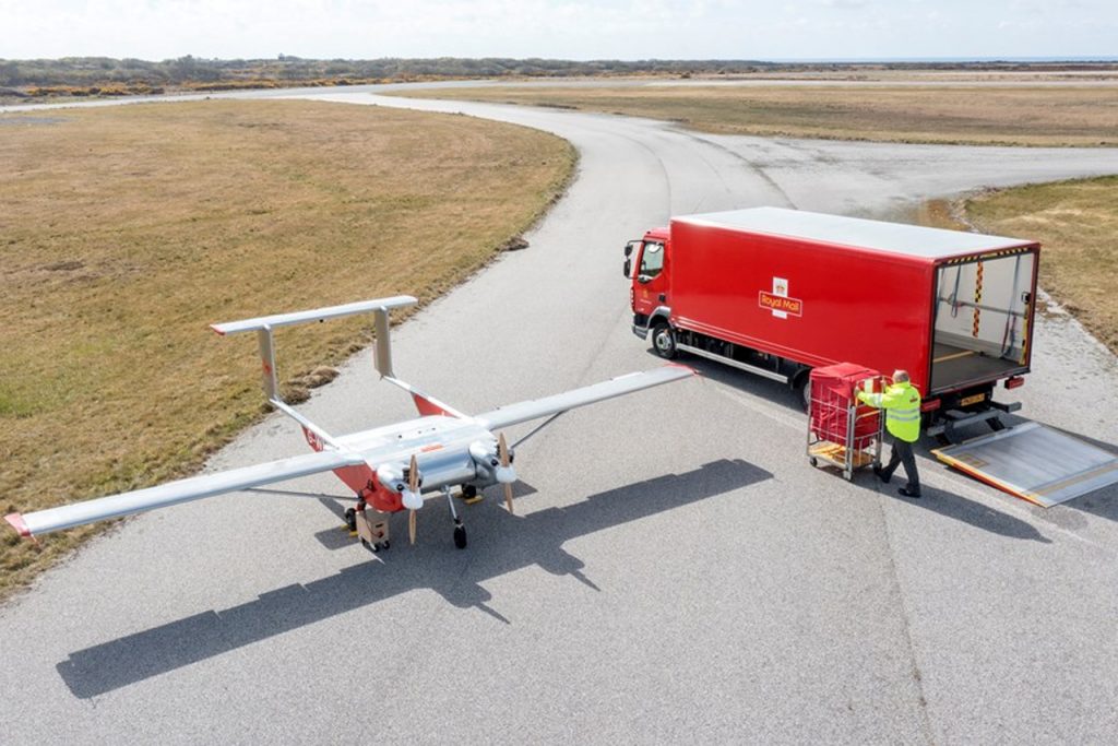 A royal mail parcel truck, a royal mail worker, and a Royal Mail autonomous cargo delivery drone stationed on a road in a field. 