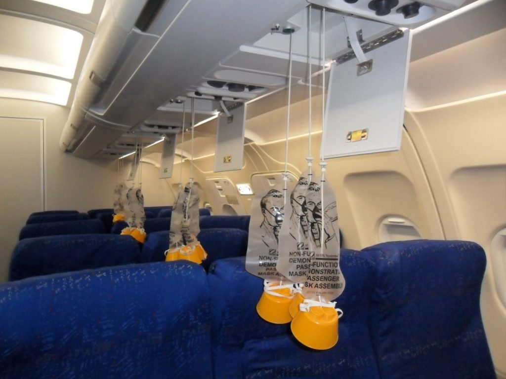 A pressurized aircraft cabin with demo oxygen masks hanging from the panel above passenger seats. 