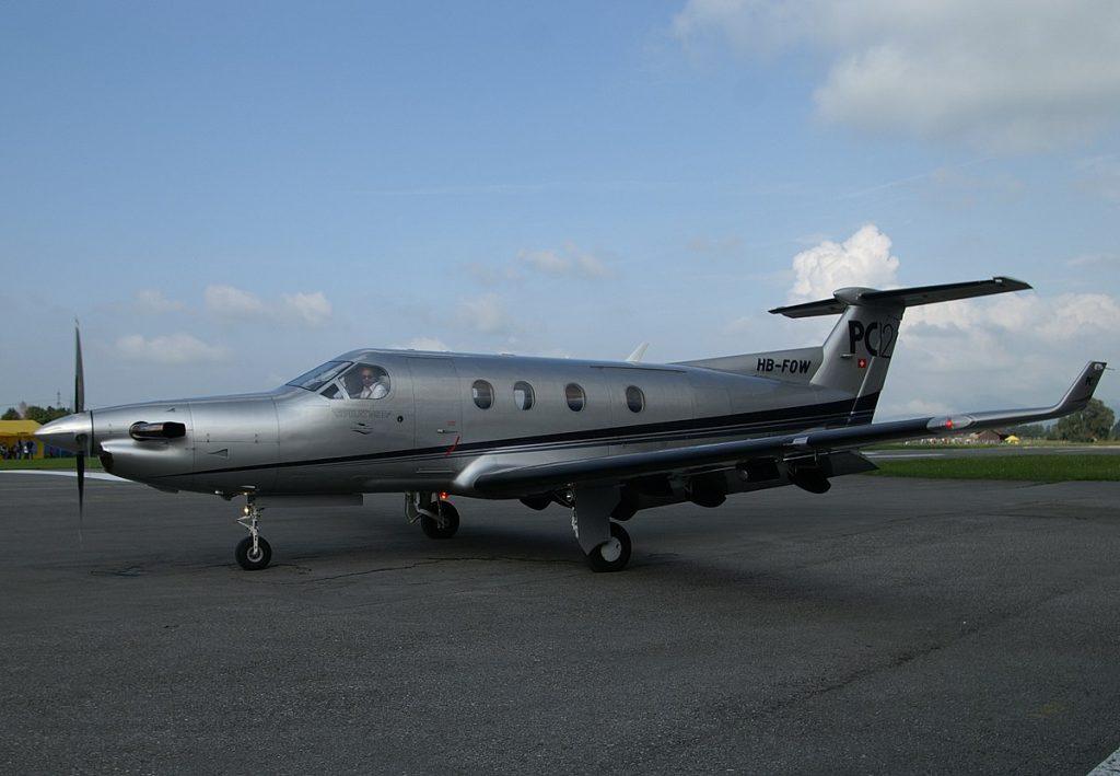 A sideways view of a silver Pilatus PC-12 with its pilot at an airfield.