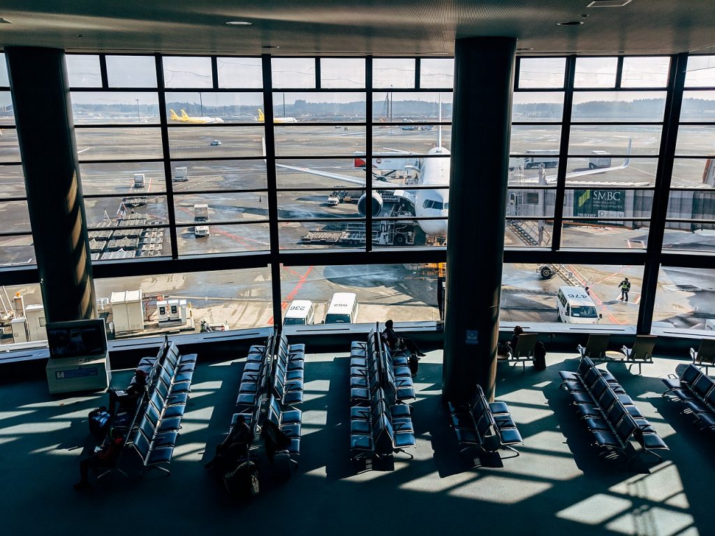 A view of an airport through a waiting hall window: an aircraft parked by a jet way and being serviced by ground staff before another flight. 