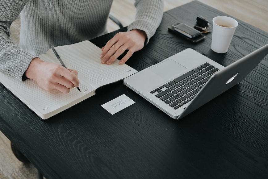 A woman sitting at a desk and taking notes in online learning courses while having a cup of coffee. 