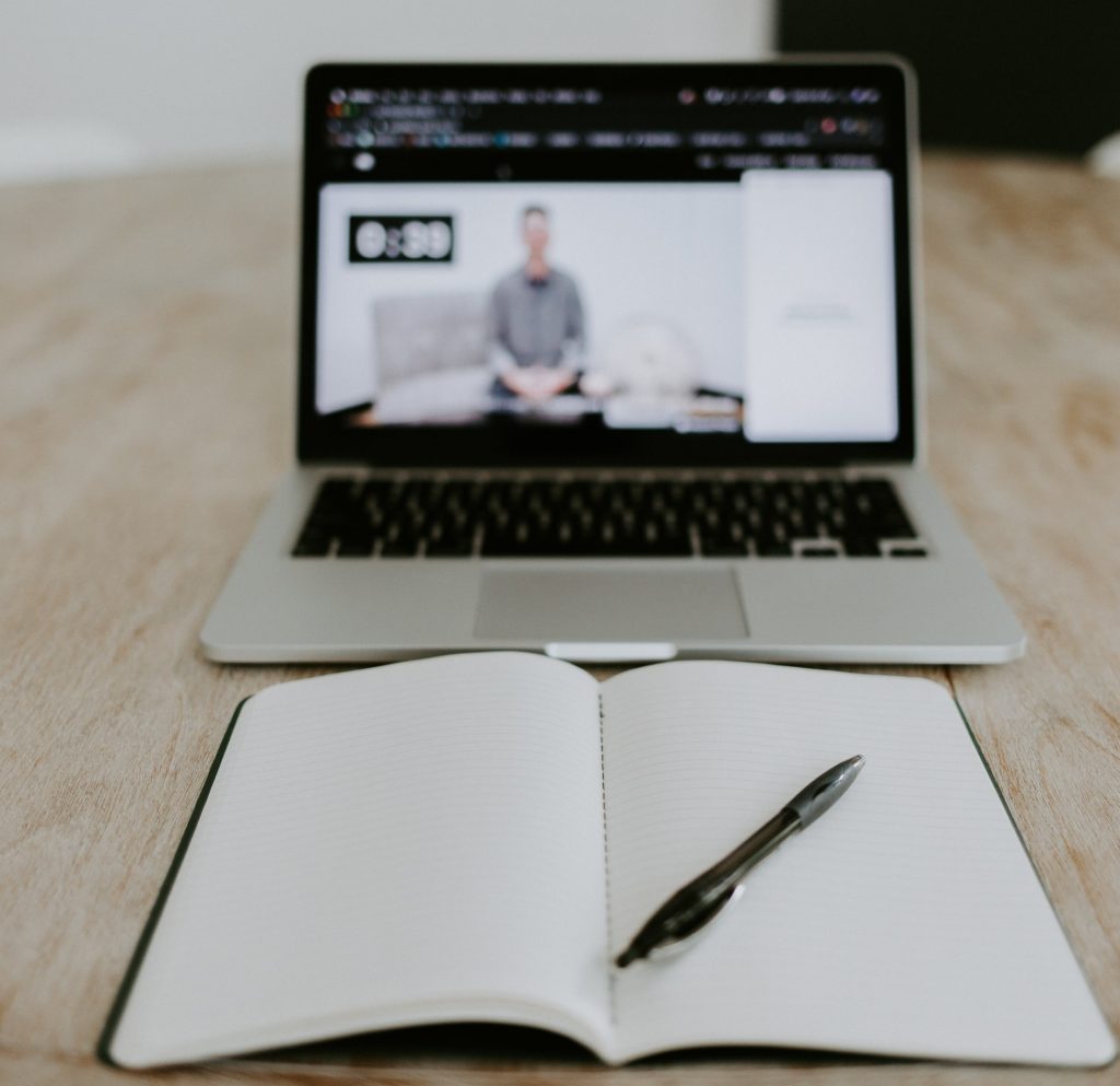 A person's, who is participating in a virtual classroom via laptop, table with an empty notebook.