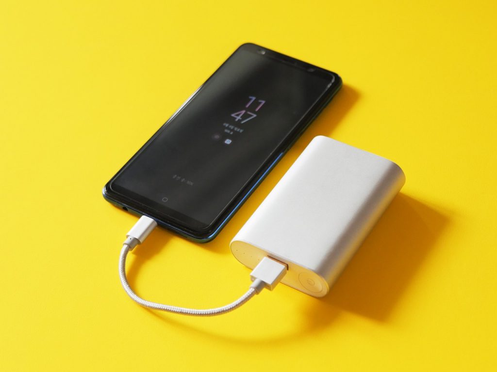 A central view on a yellow background of a black smartphone connected to a white portable charger.