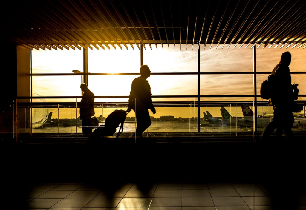 Travelers with luggage walking down a windowed airport terminal with a view of airplanes in the evening.