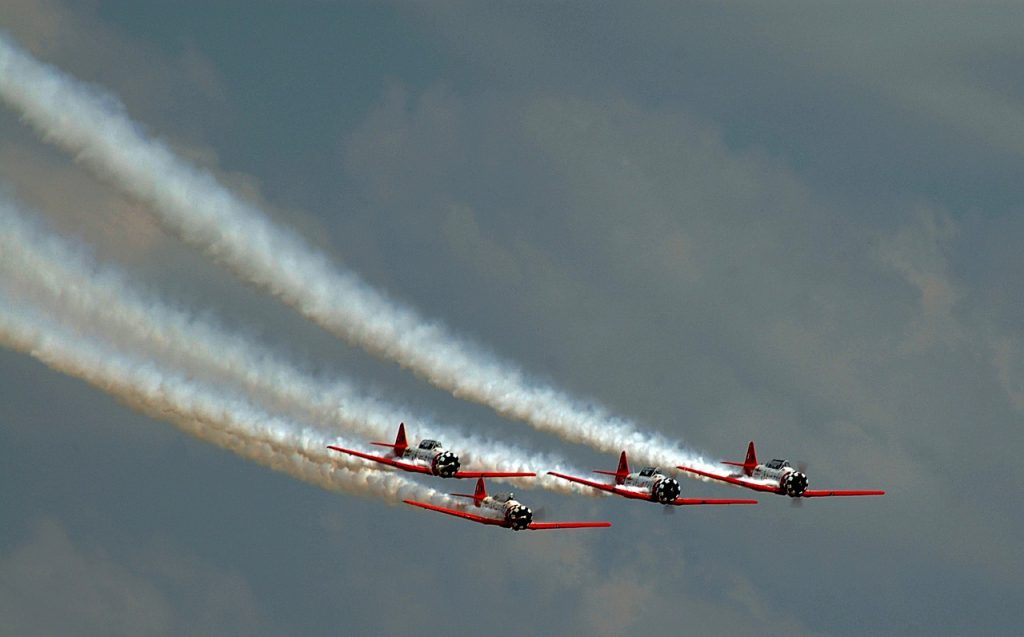 Four orange and white stunt planes in an airshow in flight across the sky with a vapor trail behind them.