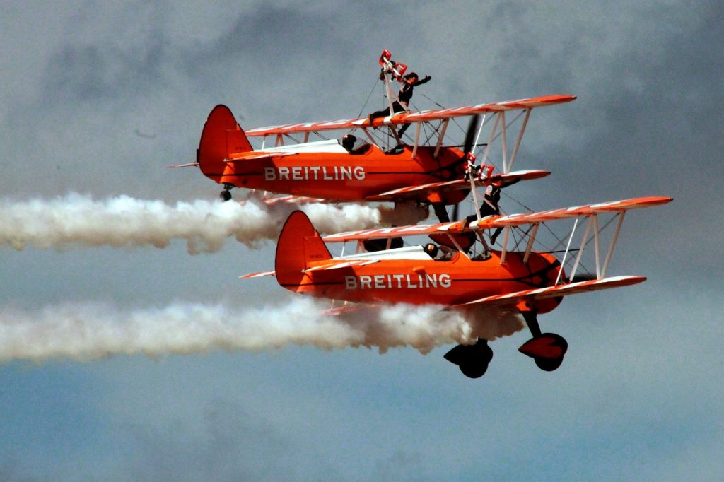 Two orange biplanes leaving a vapor trail with their pilots on wings, performing a stunt.