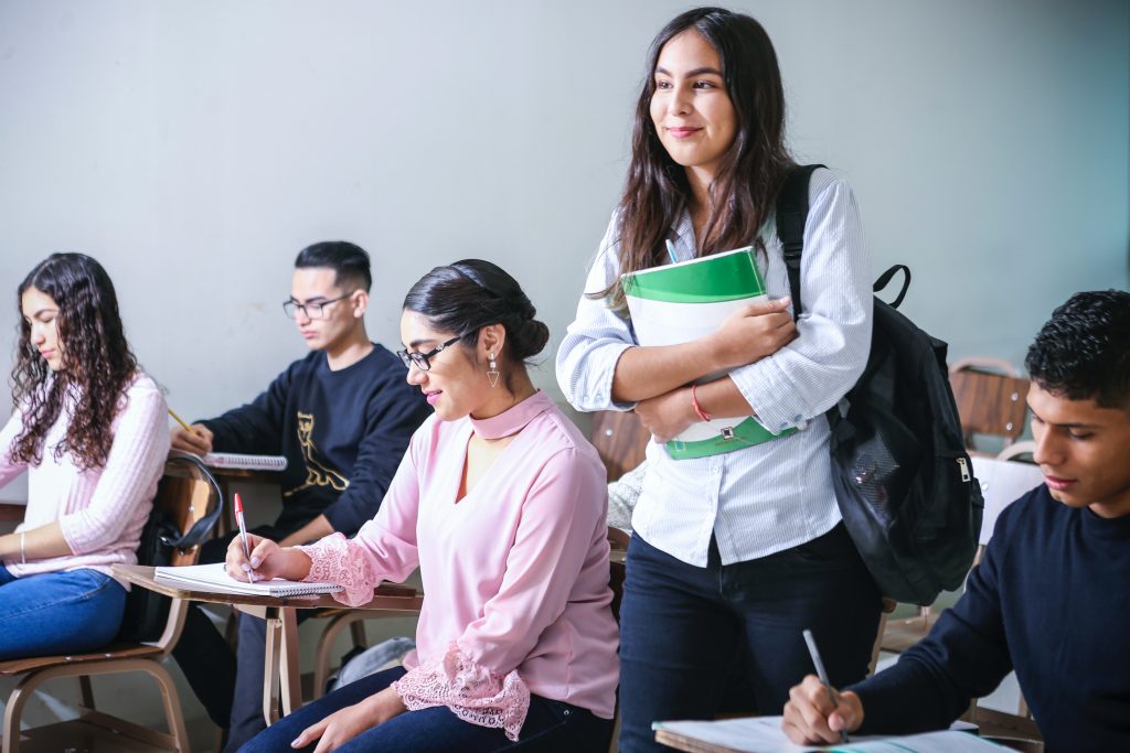 A female student is standing in the middle of the class holding books and papers. 