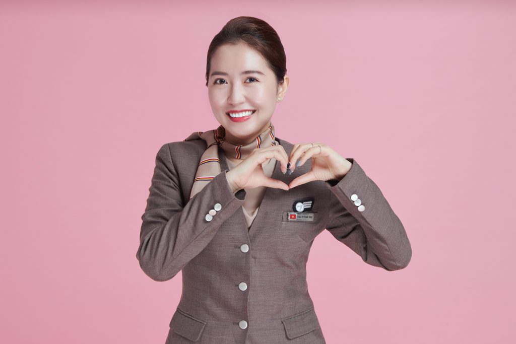 A Vietnamese flight attendant standing in front of a pink background in her uniform.