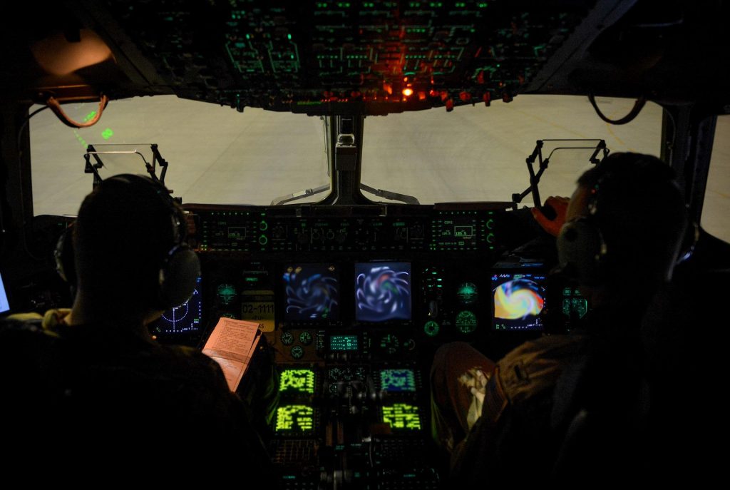 Two pilots are sitting in a dark cockpit illuminated by the instrument panel. 