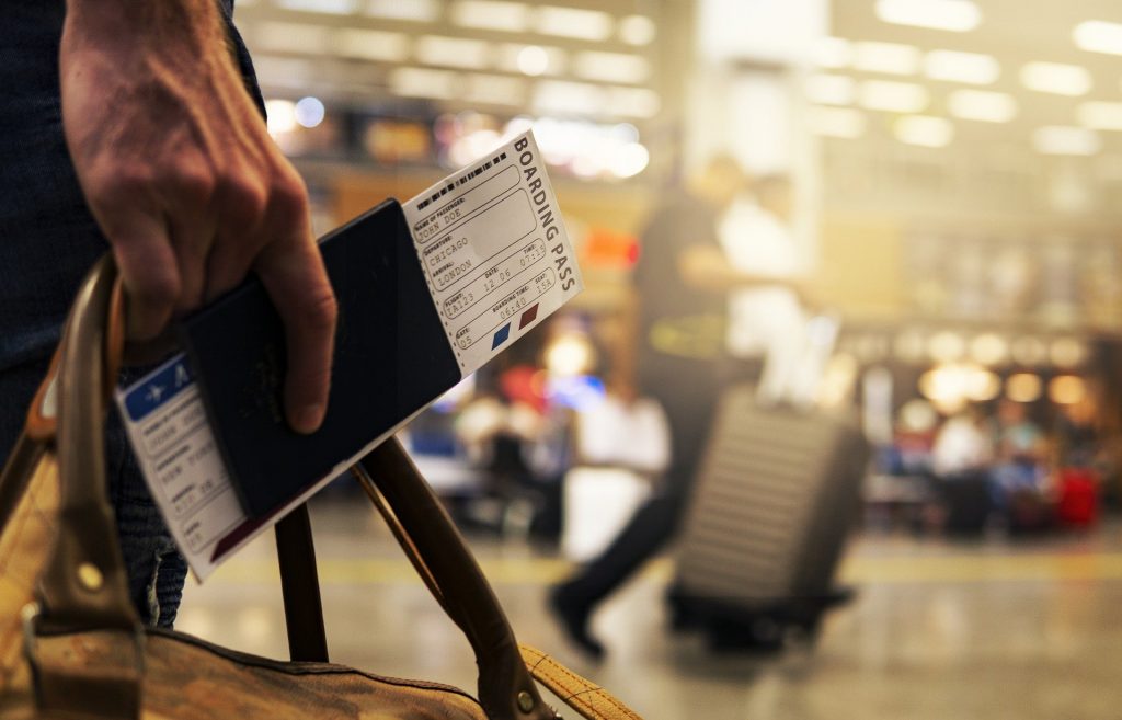 A person at the airport holding a travel bag, a passport and their boarding pass.