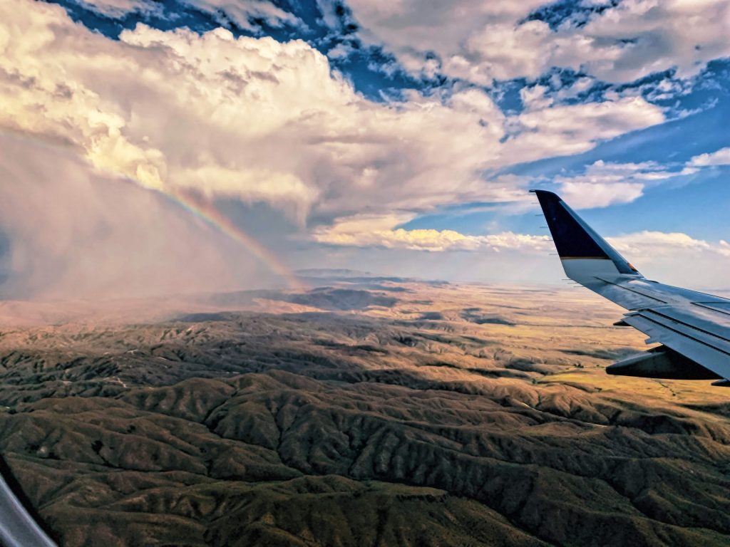 A wing of a plane over green fields and a rainbow in the distance.