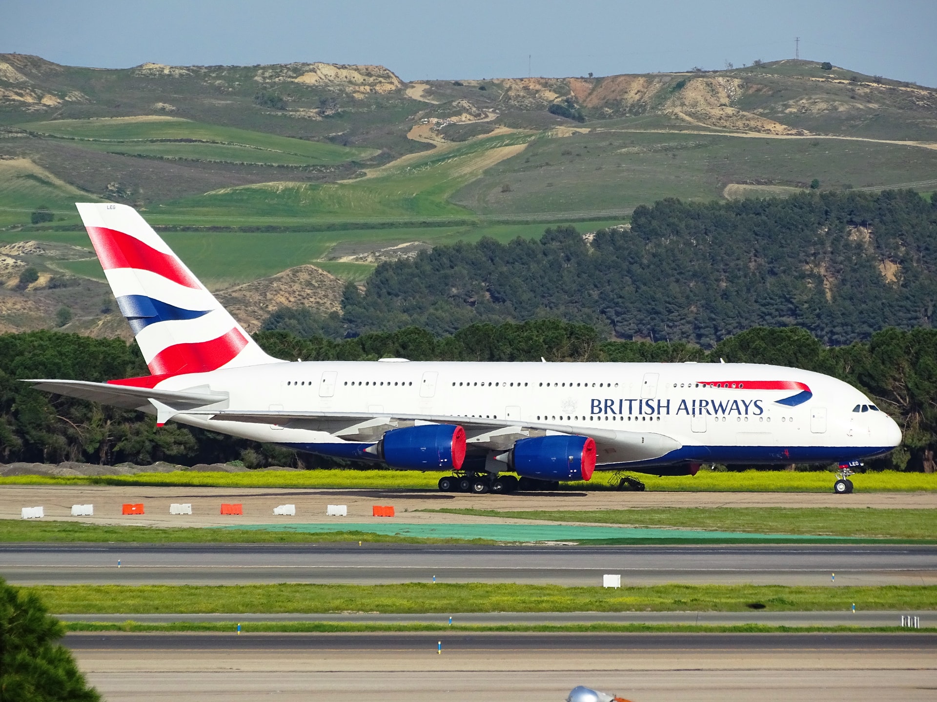 Low environmental impact British Airways aircraft landing on a runway next to green fields and mountains.